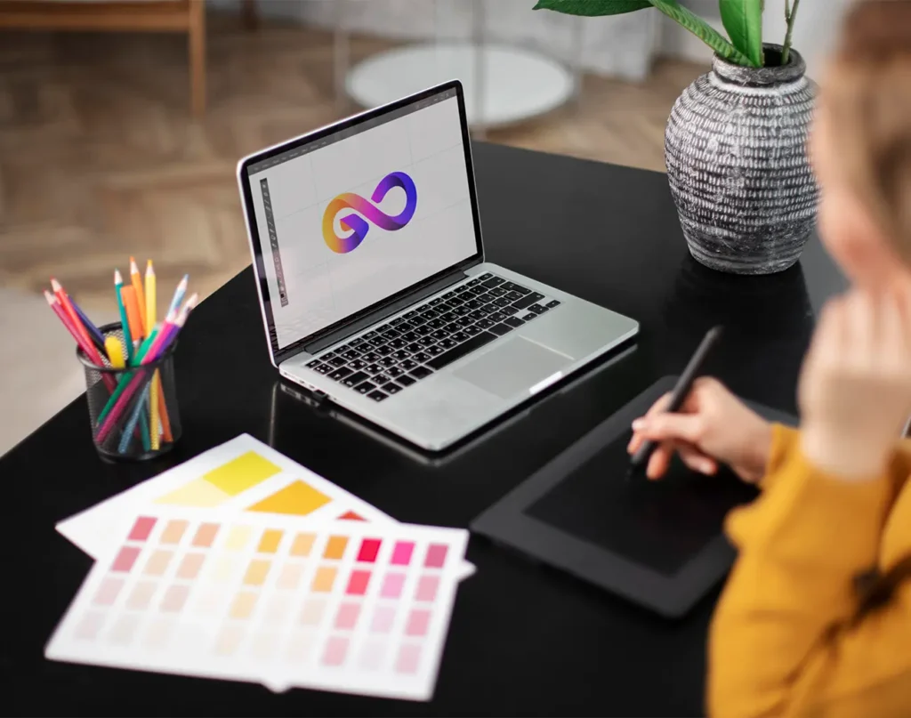 A person using a drawing tablet, with a laptop showing an infinity logo, surrounded by color swatches and art tools.