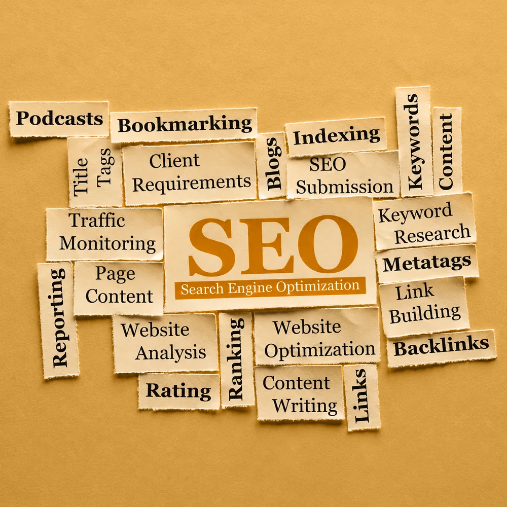 Scattered paper strips printed with SEO-related terms like "Keywords," "Backlinks," and "Traffic Monitoring" surround a central card with the large acronym "SEO" on a textured yellow background.