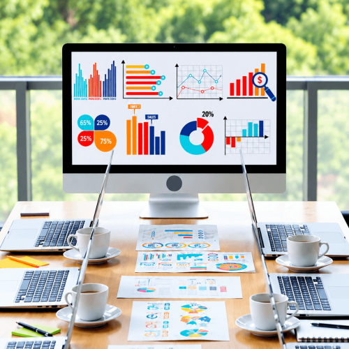 A large monitor displays colorful charts and graphs above a table covered with laptops, printed charts, and coffee cups; “SALES” and “20%” are visible.