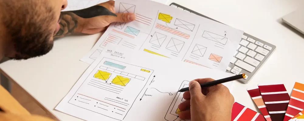 A designer sketching a website wireframe on paper, surrounded by color swatches and a keyboard, focusing on UI/UX design and branding.