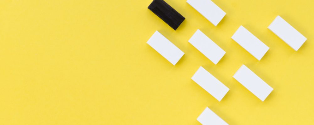 A black domino stands out among a formation of white dominoes on a bright yellow background.