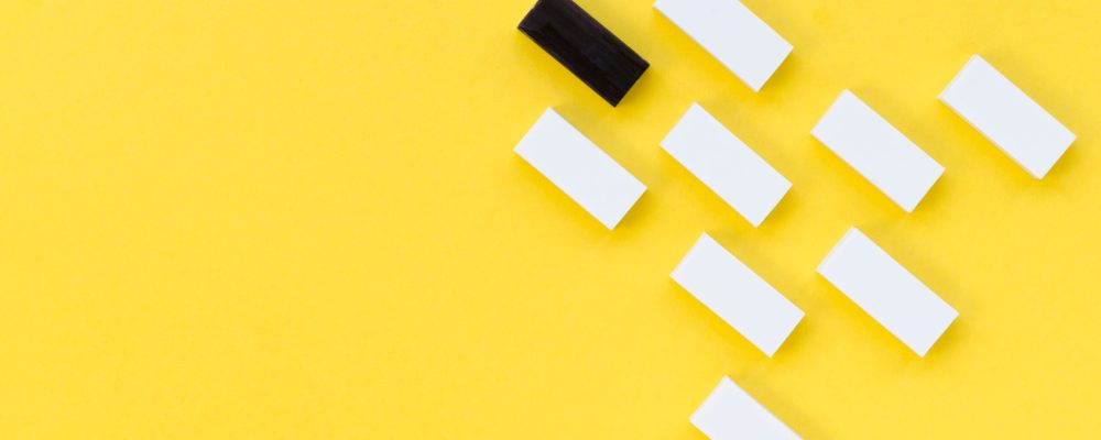 A black domino stands out among a formation of white dominoes on a bright yellow background.